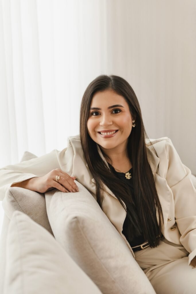 Médica psiquiatra sentada em sofá, sorrindo, vestindo roupa elegante, transmitindo postura profissional, confiança e acolhimento em ambiente tranquilo.