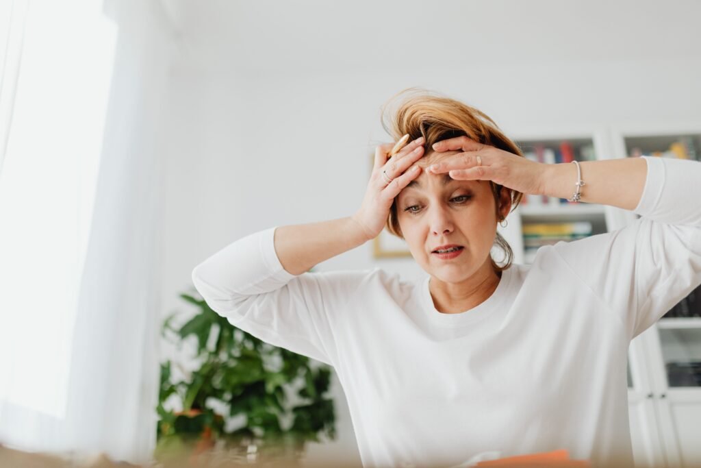 Mulher em ambiente interno, com expressão de tensão e a mão apoiada na cabeça, transmitindo sensação de sobrecarga mental. A imagem ilustra estados de ansiedade e estresse, frequentemente associados a preocupação intensa, fadiga emocional e dificuldade de relaxamento.